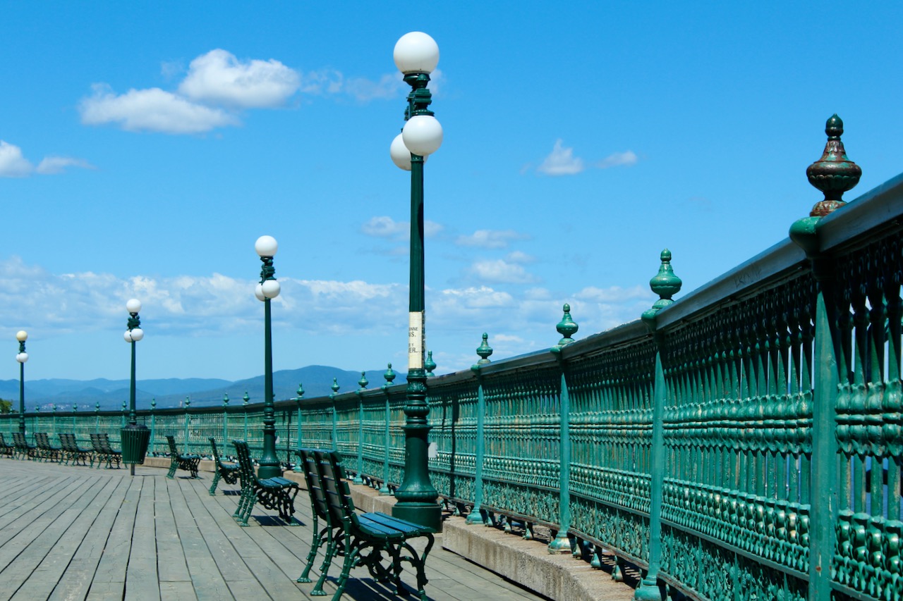 An outdoor promenade with a wooden boardwalk, decorative railing, and streetlamps. A sunny day with a blue sky in the background